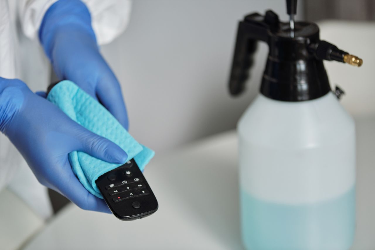 Gros plan sur des mains gantées désinfectant une télécommande avec un chiffon et un flacon pulvérisateur.